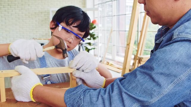 Happy Asian father and son work as a woodworker and carpenter. Father teaching his son to hammer nails on a wooden plank carefully together. carpentry working at a home workshop studio.