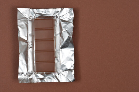 Milk chocolate bar placed on an open silver aluminum foil wrapper on a brown background with copy space on the right