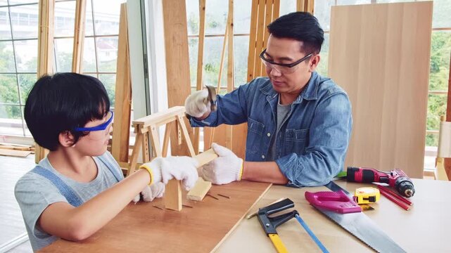 Happy Asian father and son work as a woodworker and carpenter. Father teaching his son to hammer nails on a wooden plank carefully together. carpentry working at a home workshop studio.