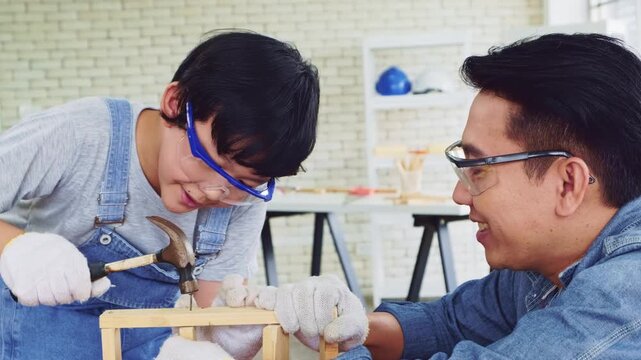 Happy Asian father and son work as a woodworker and carpenter. Father teaching his son to hammer nails on a wooden plank carefully together. carpentry working at a home workshop studio.