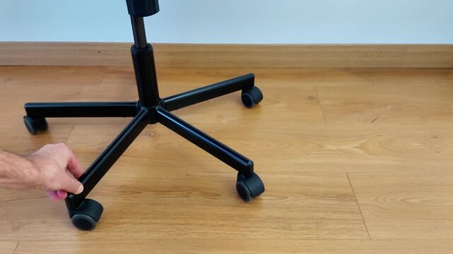 A hand grips the caster wheel at the base of a black office chair on a wooden floor, showing a clear moment of adjustment or maintenance against a clean indoor background