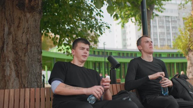 two friends sitting under tree on bench in reflective mood, closeup portraits show thoughtful gazes and slow sips from bottle, relaxed posture, urban park backdrop with foliage and soft bokeh, calm