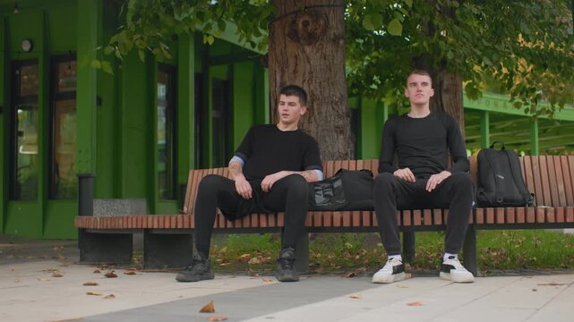 white young men waiting on bench by green pavilion, one stares skyward while other checks phone, backpacks at side, fallen leaves on pavement, tense silence and subtle distance between companions,