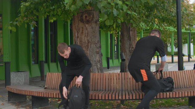 two men rummaging through backpack on park bench near green pavilion, one retrieving bottle and handing to friend, cooperative gesture moves from urgency to relief, campus sidewalk setting, supportive