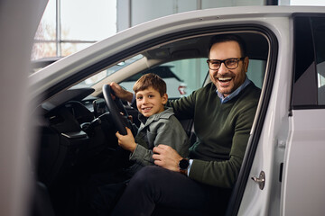 Happy father and son testing new car in auto salon and looking at camera.