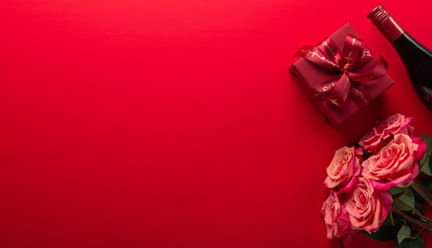 Gift, present box with red ribbon bow, rose flowers and wine bottle on red background top view. Valentine's day, 14 February, birthday, 8 march women's day gift. Love and romance.