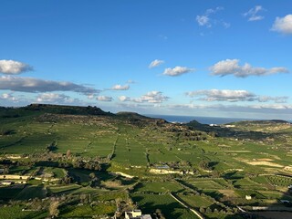 Wide rural landscape with green fields, rolling hills and blue sky, peaceful countryside scenery