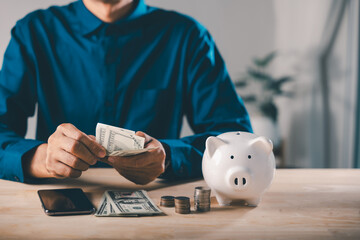 Man counts cash for digital savings mobile payment bank, piggy bank and phone help save online using smartphone pig app, holding dollar bills near stacks of coins on wooden table