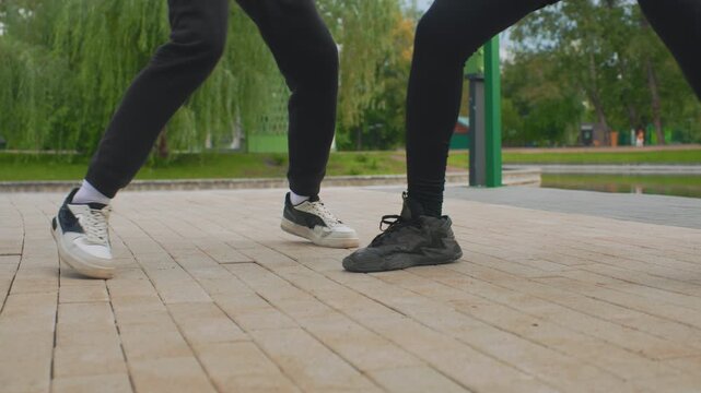 park pavement closeup dancing feet perform rapid shuffle duel on tiled promenade, two young dancers in contrasting sneakers execute precise tapping and footwork, willow trees and green posts frame
