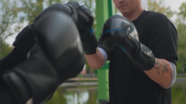 outdoor boxing mitt work white boxer training with coach pads by lake, black gloves, tattooed forearm, focused stance, crisp jabs and hooks, green pullup bars in background, morning light, informal