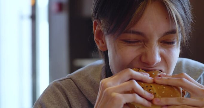 In a cozy cafe, a Caucasian teenage girl happily eats a burger, savoring the delicious flavors and enjoying a peaceful moment away from the busy world.