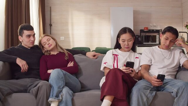 Young multiethnic friends couple sitting sofa with mobile cell phones, distracted cellphones during social gathering, digital addiction, disconnected interaction. Modern communication problem issue.