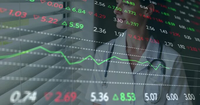 Female medical expert jotting notes on clipboard by blinds as financial charts scrolling for review