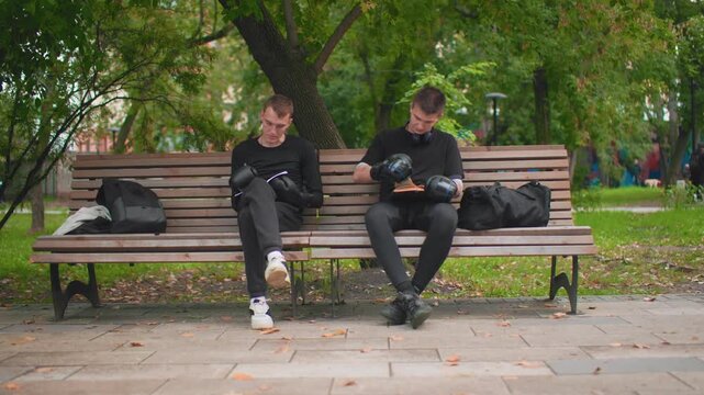 Two white men sharing boxing gloves on bench, exchange of gear, brief conversation, relaxed urban park setting, backpacks and duffel nearby, sidewalk and fallen leaves, passerby in background, mutual