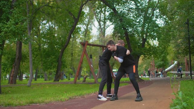 Two Caucasian men in park playfully exchanging prank and apology near swings, lighthearted carry and laugh, handshake and hug follow, casual black outfits and sneakers, afternoon greenery