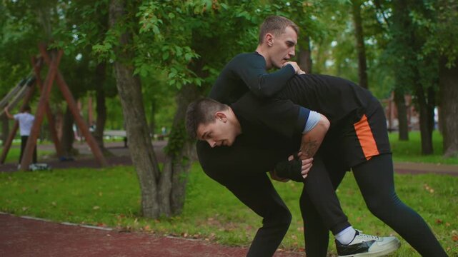 white male coach holding pads in park, focused mitt training under trees, student throwing measured punches, clinch and takedown practice, gloves, sweat, determination, evening green backdrop, urban