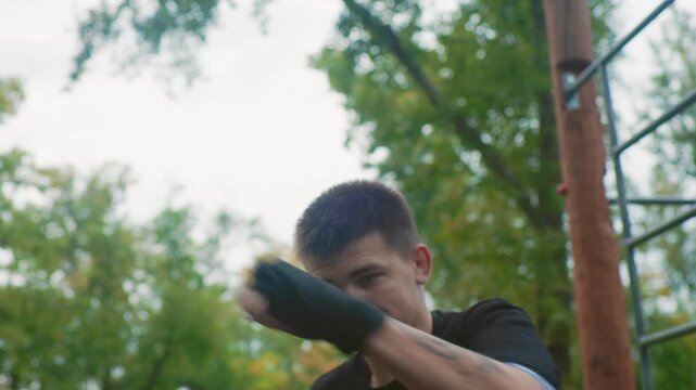 white man shadowboxing near wooden bars delivering rapid punch combinations and defensive head movement, wearing black hand wraps and training shirt against leafy overcast backdrop, closeup facial