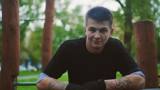 white male smiling at pullup bar in leafy park, approachable coach vibe with tattoos and gloves, engaging eye contact, leaning on rail while chatting, warm evening light and community energy, candid