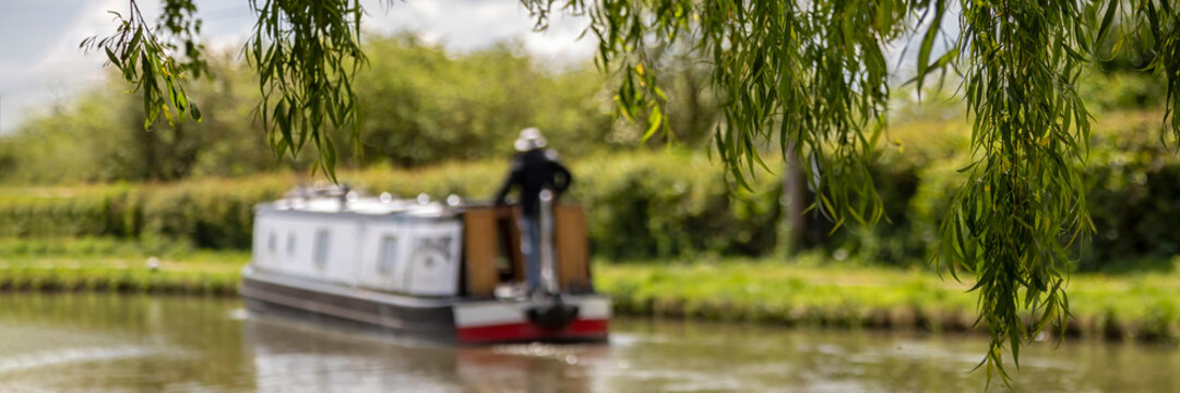 Panorama of defocused narrowboat on a canal seen through the leaves of a weeping willow