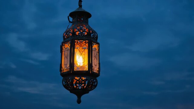 A single hanging lamp illuminates the darkness of a dusky evening. The lamp showcases intricate designs and warm light, casting shadows on the nearby surface.