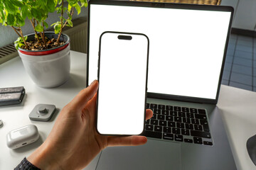 POV top view minimal desk setup mockup. Hand holding phone with copy space, laptop, earphone and a...