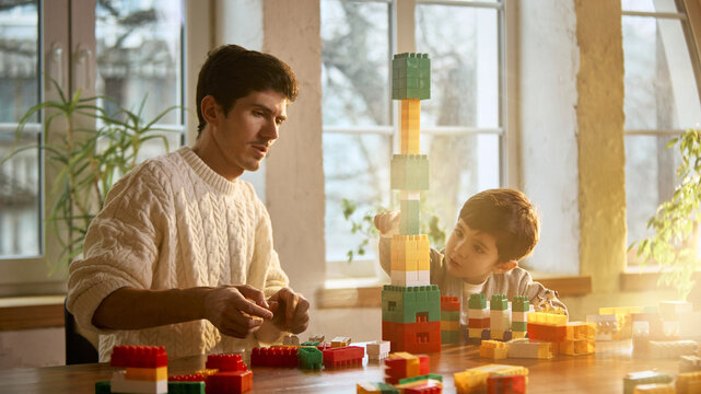 Father and child creating tall block tower together at home. Concept of teamwork, creativity growth, shared achievement, learning through play and strong family connection.