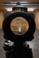 Point of view through a telescopic rifle scope with red illuminated reticle aiming at a target in an indoor shooting range.