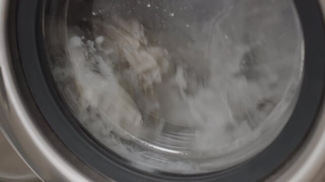 Close up transparent washer door showing soapy water and fabric tumbling inside, droplets streaking glass, stainless rim in frame,clean household textiles swirling under foam