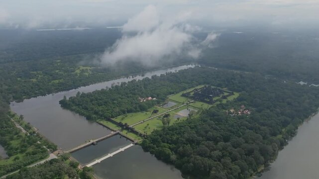 Aerial drone wide angle view showing Bakong temple complex surrounded by rectangular moat, dense forest, waterways, and green landscape under soft daylight in Cambodia.