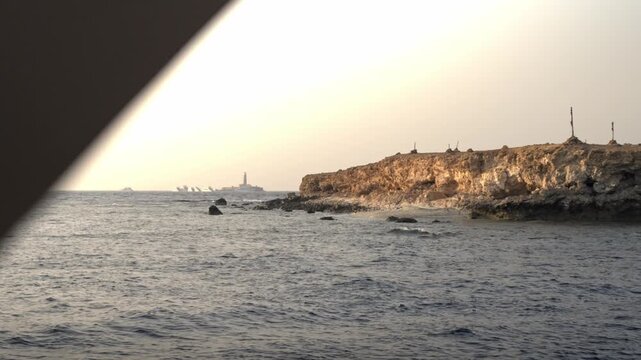 Little Brother Island during sunset. The rocky, arid cliffs of the island are illuminated by the warm, fading light, casting golden tones over the stone and the dark, choppy waters of the Red Sea