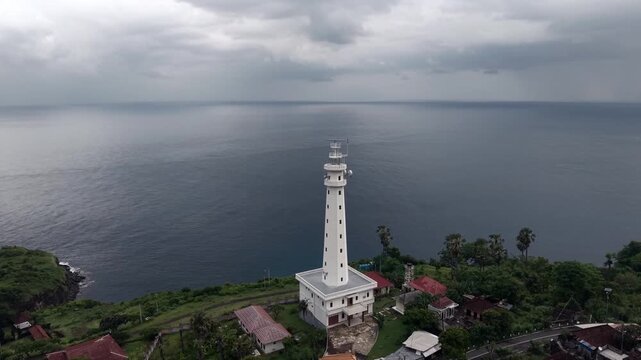 Aerial drone view of white lighthouse on tropical cliff overlooking the ocean, dramatic cloudy sky, coastal landscape in Indonesia.
