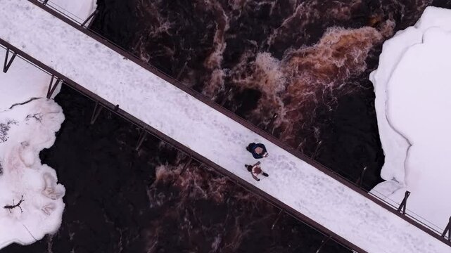A couple walking over a snow bridge above reddish brown water river, Finland, Aerial