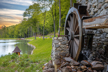 An old wooden water wheel attached to a stone wall turns under flowing water next to a peaceful lake surrounded by lush green forest.