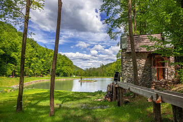 An old wooden water wheel attached to a stone wall turns under flowing water next to a peaceful lake surrounded by lush green forest.