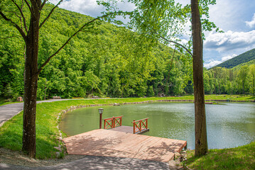 A wooden pier sits on the edge of a calm green lake surrounded by lush forested mountains under a cloudy sky.