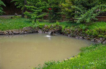 A small wooden water flume pours a steady stream into a murky pond surrounded by lush green foliage and stone banks in a park setting.