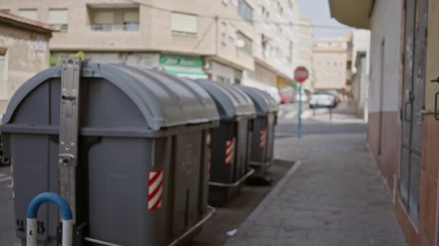 Row of gray trash containers lined along a narrow sidewalk in soft blurred street background; background backplate copyspace calm.