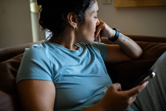 Thoughtful woman holding smartphone at home on couch