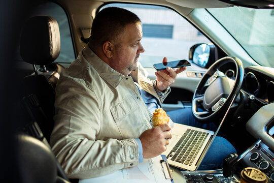 Businessman multitasking with phone and laptop while eating in car