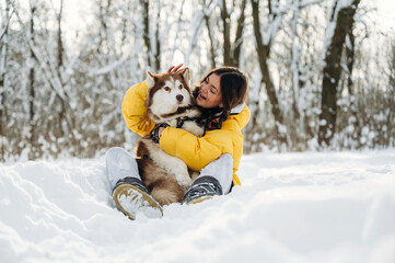 Obraz premium Young woman in a yellow puffer jacket is sitting in the snow while holding a brown and white husky dog, surrounded by snow-covered trees in a winter landscape