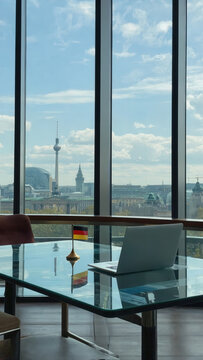 A modern office with a laptop on a glass table. A small German flag is displayed. The view includes a city skyline with tall buildings and a tower.