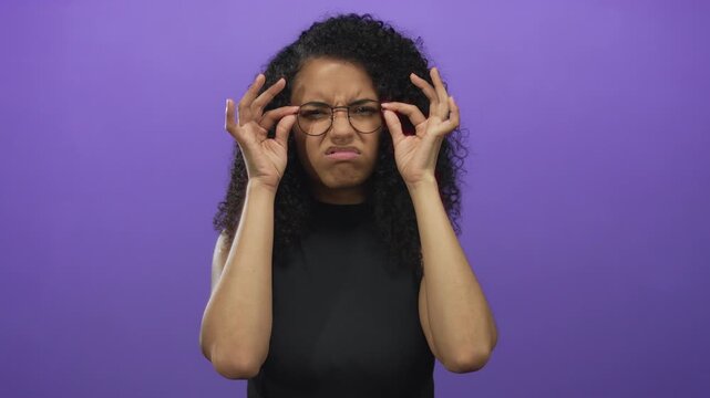 Young woman adjusting glasses with a confused expression against an isolated purple background showcasing her curly hair and black shirt