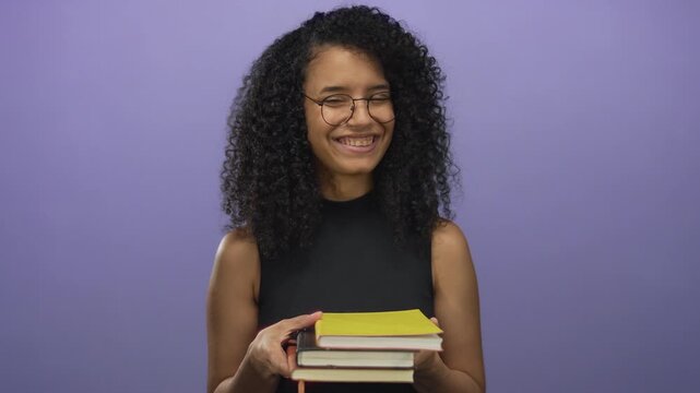 Young hispanic woman smiling and holding books against an isolated purple background, showcasing a vibrant and studious atmosphere.