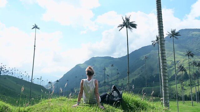 Back view of a young woman sitting on a gentle green hillside with her backpack resting on the grass, quietly observing the breathtaking Cocora Valley in Salento, Colombia. The iconic tall wax palms 