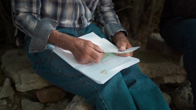 Close-up of adult man in plaid shirt sitting outdoors, sorting papers and mail on his lap&mdash;bills, paperwork review, personal finance and decision-making moment.