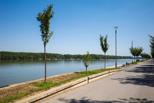 Danube promenade Vukovar, Croatia. Walkway lined with trees and streetlights, offering views of the river and the forested opposite bank. In the background, a clear blue sky.