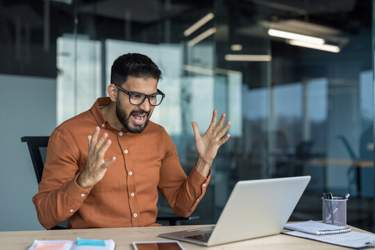 Young bearded businessman expressing anger and frustration while looking at a laptop screen in a modern office, reacting to a software problem or bad news