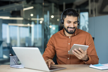Young indian man wearing a headset and smiling, sitting at a desk and simultaneously using a laptop and a digital tablet, working in a modern office environment