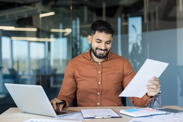 Young confident bearded man holding documents and typing on laptop, reviewing paperwork and analyzing data while sitting at a desk in a contemporary clear glass office environment