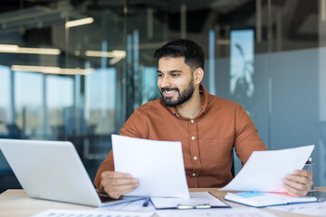 Young indian businessman at a modern office desk, smiling while reviewing reports and laptop,...